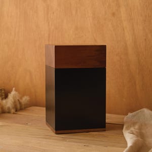 May include: A rectangular wooden box with a dark brown top and a black body. The box is sitting on a wooden surface. The background is a light brown wooden wall.