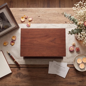 May include: A wooden memorial box with a dark finish sits on a wooden table, next to a framed black and white photo. A guest book, pen, and lit candles are also on the table, along with dried flowers and petals.