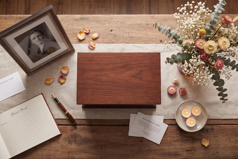May include: A wooden memorial box with a dark finish sits on a wooden table, next to a framed black and white photo. A guest book, pen, and lit candles are also on the table, along with dried flowers and petals.