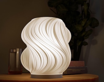 May include: A white, sculptural table lamp with a unique, flowing design. The lamp's shade features a series of curved, vertical lines, creating a wave-like pattern. It sits on a round, dark base and is placed on a wooden table.