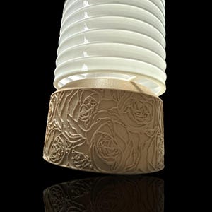 May include: A white lampshade with a brown base featuring a rose pattern. The lampshade is made of a ribbed material and has a cylindrical shape.