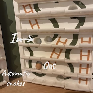 以下が含まれることがあります： 複数のレベルと一連の穴がある白い木製ゲーム。ゲームには「Automatic snakes」、「In」、「Out」のテキストが表示されます。デザインには、緑色のヘビのような形とオレンジ色の文字「H」のパターンが含まれています。