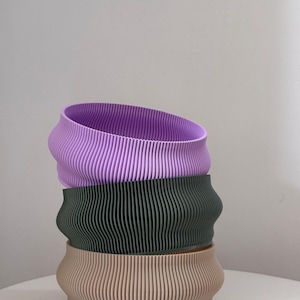 Può includere: Tre fioriere impilate e a coste nei toni lavanda, verde oliva e beige. Le fioriere hanno una forma ondulata e organica e sono realizzate in un materiale solido. Il design presenta linee verticali che creano una superficie strutturata. Queste fioriere decorative sono adatte per interni.