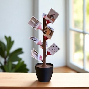 Könnte beinhalten: Ein brauner Foto-Baum-Display mit schwarzer Topfbasis. Der Baum hat rosa und rote Clips, die mehrere kleine Fotos halten. Das Display steht auf einer Holzoberfläche in der Nähe eines Fensters.