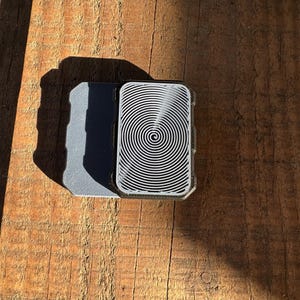 May include: A gray and black rectangular case with a spiral pattern on the front.