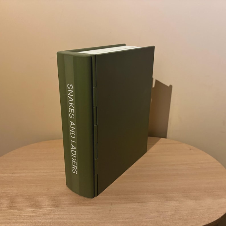 以下が含まれることがあります： 木製のテーブルの上に置かれた、緑色のボードゲーム「Snakes and Ladders」。ゲームの背表紙には白い文字でタイトルが表示されています。ゲームのカバーは、くすんだ緑色です。