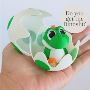 Op de afbeelding: Een groene dinosaurusfiguur in een witte eierschaal, vastgehouden in een hand. De dinosaurus heeft grote zwarte ogen en oranje details. De eierschaal is versierd met groene stippen. De tekst op de afbeelding luidt: "Do you get the Dinoshi?"