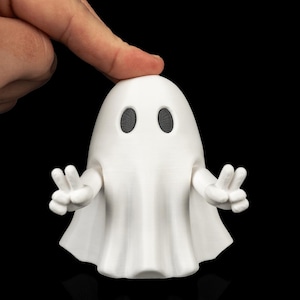 Op de afbeelding: Een wit 3D-geprint spookfiguurtje met een gladde, ronde kop en twee ovale zwarte ogen. Het spook heeft twee beweegbare armen die naar buiten zijn gestrekt, waarbij elke hand een vredesteken vormt met zichtbare lijnen van de 3D-printstructuur.