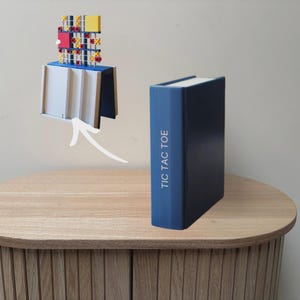 May include: A blue book-shaped box with the text TIC TAC TOE printed on the spine. Above the box, a white arrow points to a game board featuring a grid with red and yellow X and O markers, set on a white and blue base structure.