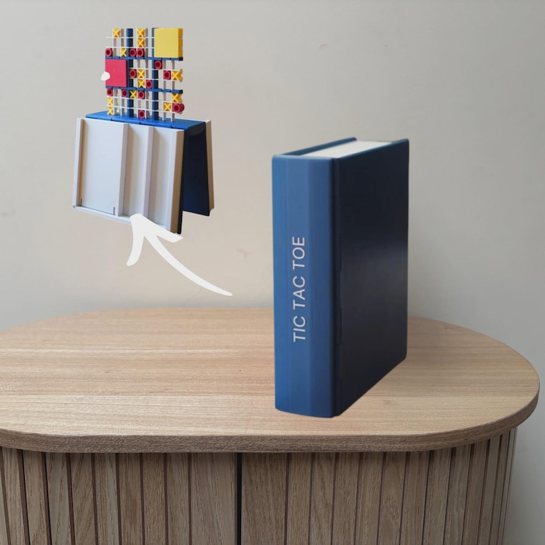 May include: A blue book-shaped box with the text TIC TAC TOE printed on the spine. Above the box, a white arrow points to a game board featuring a grid with red and yellow X and O markers, set on a white and blue base structure.