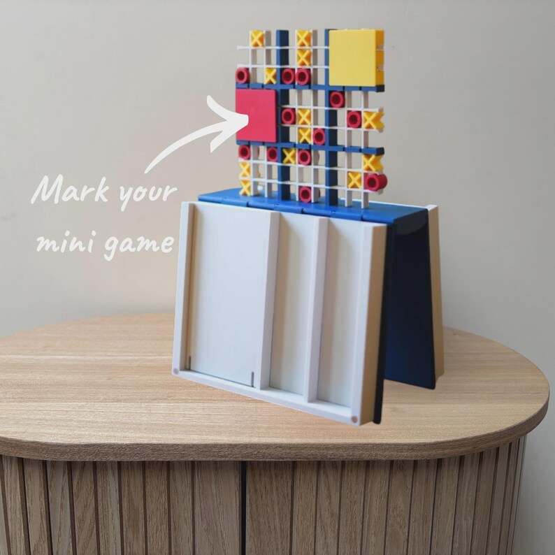 May include: A plastic tic-tac-toe game set featuring a white base with vertical slots and a blue top section holding a grid with yellow X and red O markers. A white arrow points to the grid with the text Mark your mini game printed in white.