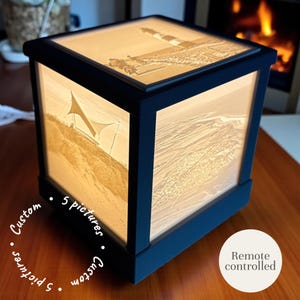 Op de afbeelding: Een zwarte, kubusvormige lamp met verlichte panelen met strandscènes en een vuurtoren. De lamp heeft een houten frame en de tekst "5 pictures" en "Custom" zijn zichtbaar. De lamp is op afstand bedienbaar.