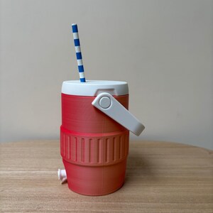 May include: A red and white cooler-style cup with a white handle and a blue and white striped straw. The cup has a white lid and a small spigot at the bottom. The cup is sitting on a wooden surface.