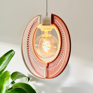 Può includere: Una moderna lampada a sospensione con una lampadina calda e luminosa. La lampada ha un design unico, con una cornice a semicerchio e un motivo intrecciato nei toni del bianco e del marrone rossiccio. Pende da un cavo bianco.