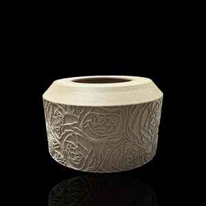 May include: A beige ceramic vase with a raised floral design. The vase has a round shape and a wide opening.