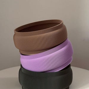 Può includere: Tre fioriere impilate e a coste nei toni del marrone, lavanda e nero. Le fioriere hanno una forma arrotondata con un design a strisce verticali strutturate. Le fioriere sono realizzate in un materiale rigido.