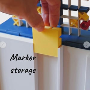 May include: A white and blue plastic game board featuring a grid for tic-tac-toe with yellow X and red O pieces. A yellow square piece is being inserted into a slot on the white base. The text Marker storage is printed in black on the white surface.