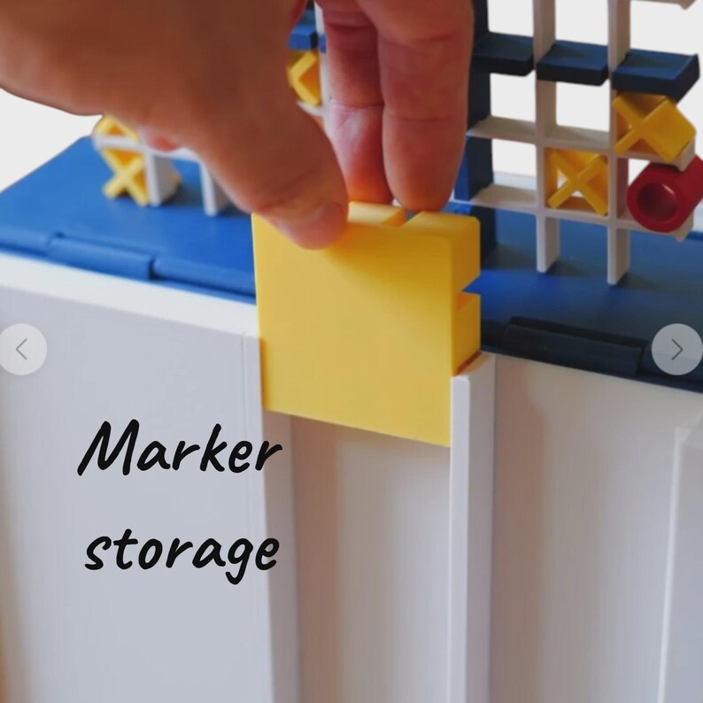 May include: A white and blue plastic game board featuring a grid for tic-tac-toe with yellow X and red O pieces. A yellow square piece is being inserted into a slot on the white base. The text Marker storage is printed in black on the white surface.