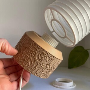 May include: A brown lampshade with a rose pattern. The lampshade is made of a textured material and has a white cord. The lampshade is being held by a hand.