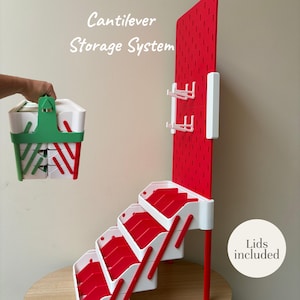 May include: A cantilever storage system featuring a red pegboard back panel with white hooks and a tiered white and red shelving unit. The text Cantilever Storage System and Lids included are visible. A separate green and white handled storage bin is shown.