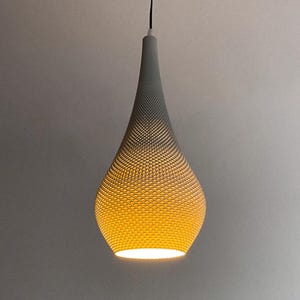 Könnte beinhalten: Eine graue, tropfenförmige Pendelleuchte. Die Leuchte hat eine strukturierte Oberfläche mit einem Muster aus kleinen, gleichmäßig verteilten Löchern. Das Licht strahlt ein warmes, gelbes Leuchten aus, das eine weiche Atmosphäre schafft.