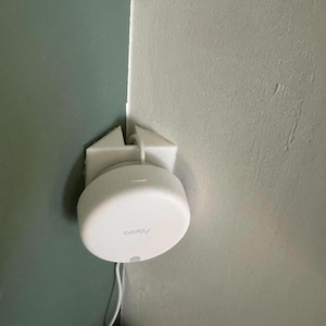 Op de afbeelding: Een witte, ronde, wandgemonteerde router met het woord "abode" erop gedrukt. De router is gemonteerd op een witte plastic beugel.