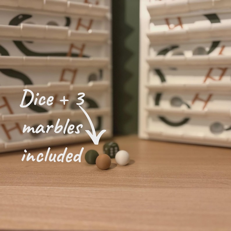 以下が含まれることがあります： 白と茶色のデザインの木製ボードゲーム。画像には「Dice + 3 marbles included」というテキストが書かれています。緑、茶色、白の3つのビー玉が木製の表面にあります。