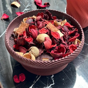 May include: A decorative, woven bowl filled with dried potpourri. The potpourri includes red, burgundy, and tan dried flowers, petals, and seed pods. The bowl is a dark mauve color and has a textured, wavy rim.