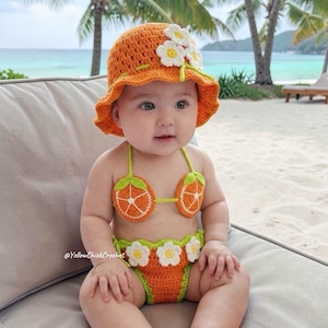May include: A baby in a handmade orange crochet sun hat and bikini set, featuring orange slice and flower details. The baby is seated on a light grey surface, with a beach and palm trees visible in the background.