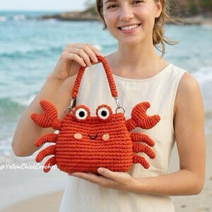 May include: A crocheted crab-shaped handbag in a vibrant orange hue. The bag features large, expressive eyes, a smiling face, and crab claws. The handle is also orange. The bag is held on a beach with the ocean in the background.