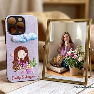 Könnte beinhalten: Eine lavendelfarbene Handyhülle aus Stoff mit einer individuellen Stickerei einer Person mit braunen Haaren, einer Katze, einer Topfpflanze und einer blauen Wolke. Der Text Emily & Jasper ist unten eingestickt. Daneben steht ein gerahmtes Foto.