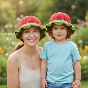 Puede incluir: Una mujer y un niño con sombreros de ganchillo a juego con temática de sandía. Los sombreros tienen una parte superior roja, un borde verde y una banda blanca. La mujer lleva una camiseta de color claro y el niño una camiseta azul y vaqueros. El fondo es un jardín borroso.