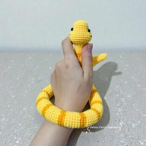 Puede incluir: Un juguete de serpiente de ganchillo amarillo y naranja hecho a mano. La serpiente está sujeta por una mano, mostrando el detallado trabajo de ganchillo. La serpiente tiene ojos negros y rayas naranjas. El juguete es un artículo suave y de felpa.