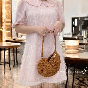 May include: A brown crochet seashell-shaped handbag with a long strap. The bag is held by a person wearing a pink dress. The bag has a label that says "Yellow Chick Crochet". The background includes tables and chairs.