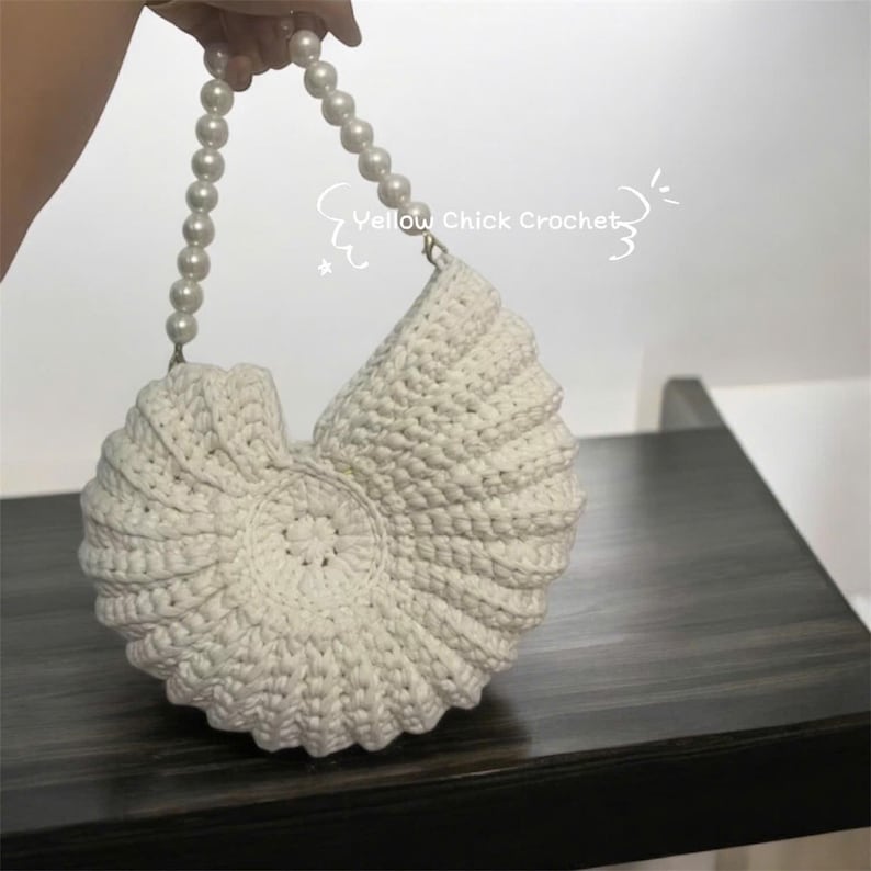 May include: A white, crochet seashell-shaped handbag with a pearl beaded handle. The bag is made of thick, textured yarn and features a spiral design. The text "Yellow Chick Crochet" is visible.