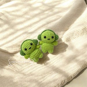 May include: Two small green crocheted plush toys with smiling faces and large ears. The toys are sitting on a white knitted blanket.