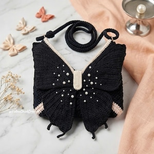 May include: A black crochet bag shaped like a butterfly with a cream-colored center and trim. The wings are decorated with small white faux pearls. The bag features a matching black crochet shoulder strap and the text @YellowChickCrochet.