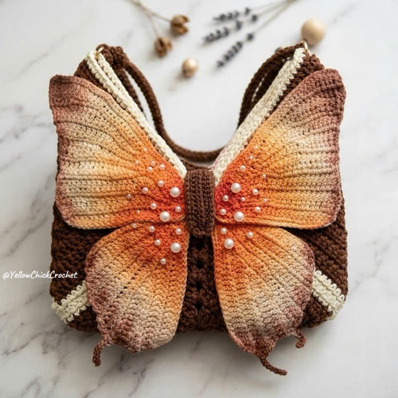 May include: A crochet handbag shaped like a butterfly with orange and brown gradient wings, featuring white pearl accents on the wings and a dark brown central body. The bag has a dark brown base and cream-colored trim, with the text YellowChickCrochet.
