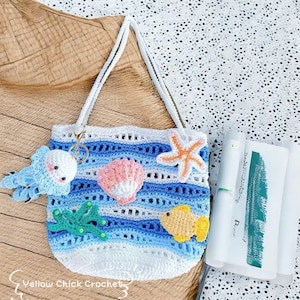 May include: A white and blue crochet tote bag with a sea-themed design. The bag features a jellyfish keychain, a pink seashell, an orange starfish, and a yellow fish. The bag has white straps and the text "Yellow Chick Crochet".