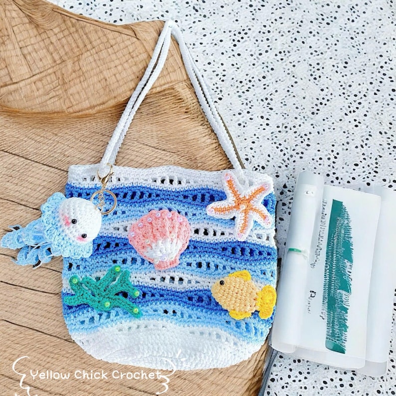May include: A white and blue crochet tote bag with a sea-themed design. The bag features a jellyfish keychain, a pink seashell, an orange starfish, and a yellow fish. The bag has white straps and the text "Yellow Chick Crochet".