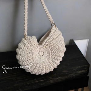 May include: Cream-colored crochet handbag with a seashell design. The bag features a rounded shape with a spiral center and a braided strap attached with gold-colored rings. The bag is sitting on a dark wooden surface.