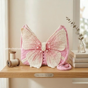 May include: A crochet shoulder bag shaped like a butterfly with pink and cream yarn, featuring a central body section adorned with small white faux pearls and a matching braided strap. The bag has a textured surface and a distinct wing design.