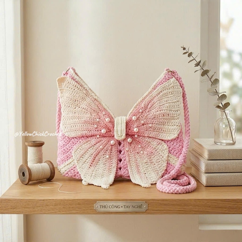 May include: A crochet shoulder bag shaped like a butterfly with pink and cream yarn, featuring a central body section adorned with small white faux pearls and a matching braided strap. The bag has a textured surface and a distinct wing design.