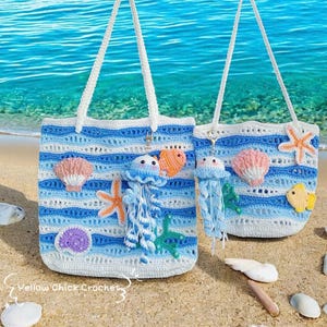 May include: Two crochet beach bags with white handles. The bags feature blue and white striped patterns, and are decorated with crochet jellyfish, starfish, seashells, and fish. The bags are on a sandy beach with the ocean in the background.