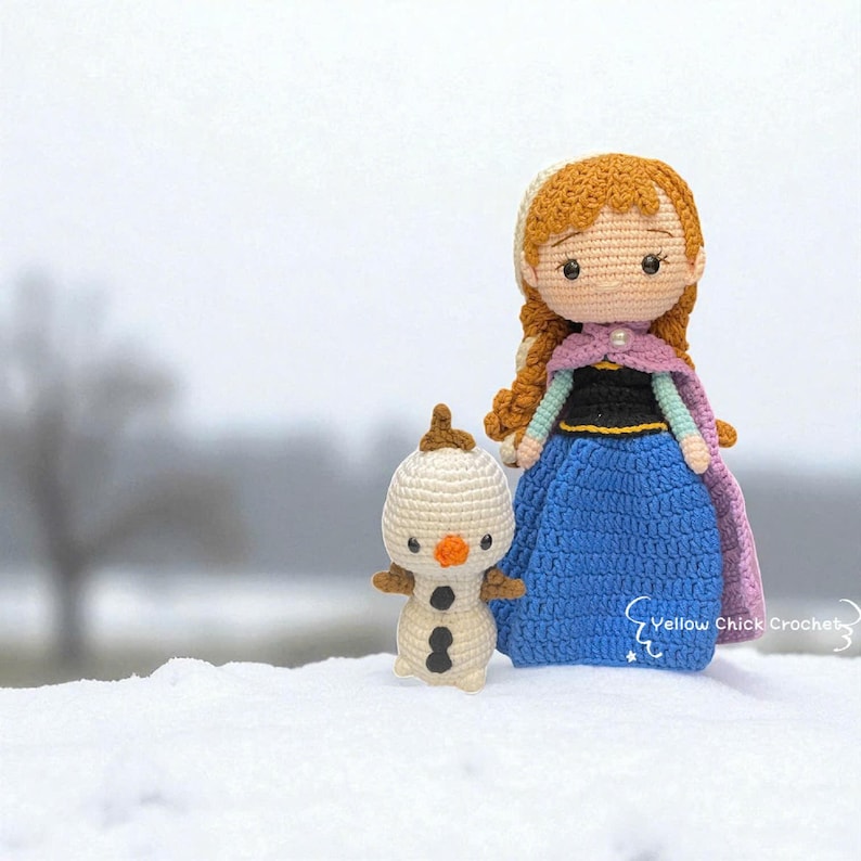 May include: Crocheted Anna doll with auburn hair, a blue dress, and a purple cape, standing next to a crocheted Olaf snowman. The dolls are handmade and set against a snowy backdrop. The Anna doll is approximately 7 inches tall.