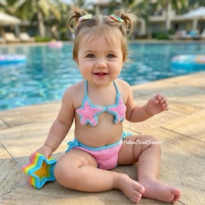 May include: A two-piece crochet bikini set featuring a pink star-shaped top with light blue trim and matching pink bottoms with light blue ties. The set includes the text YellowChickCrochet. The bikini is worn by a toddler with hair in two buns.