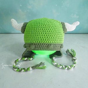 May include: A handmade crochet Viking helmet in shades of green and gray, featuring white horns and braided ties. The helmet is designed with a textured stitch pattern and includes the text "Yellow Chick Crochet".