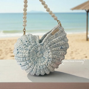 Puede incluir: Bolso de ganchillo azul claro con forma de concha, con correa de perlas. El bolso tiene un patrón texturizado y pequeñas motas azules. Una etiqueta dice "handmade". El fondo muestra una escena de playa.