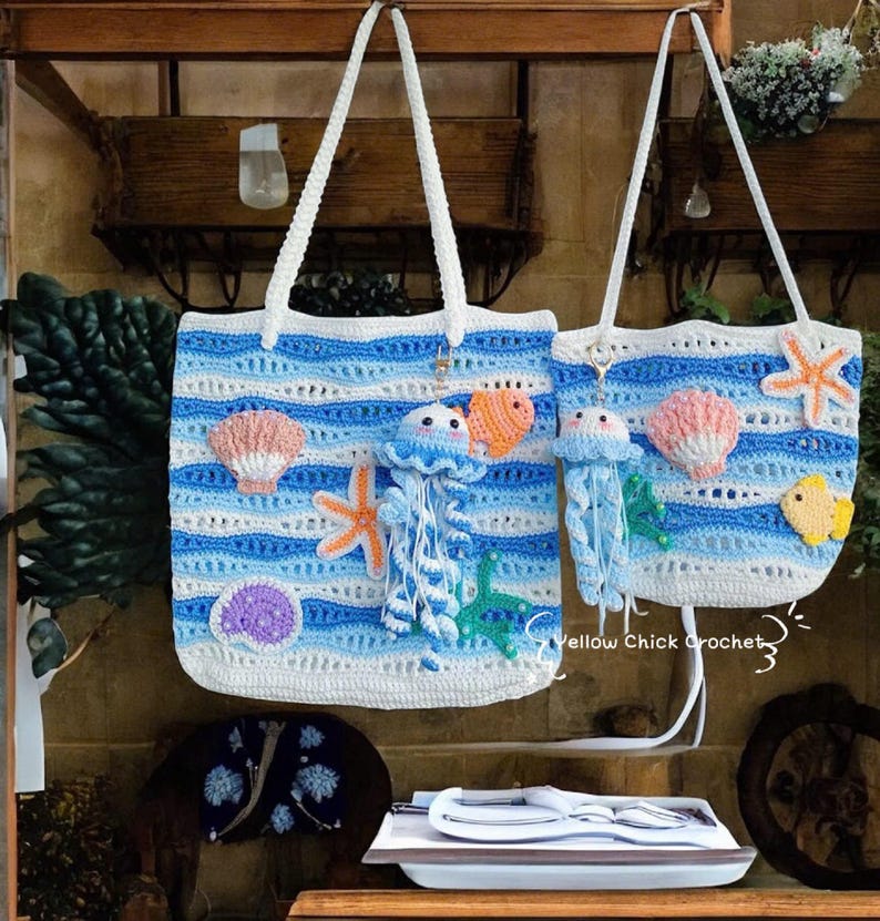 May include: Two crochet tote bags with white handles. The bags feature a blue and white striped pattern with various sea-themed appliqu&eacute;s, including jellyfish, starfish, seashells, and fish. The bags are handmade and have a beachy aesthetic.