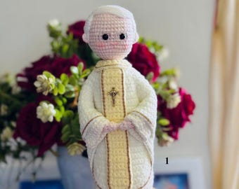 Może przedstawiać: Szydełkowana lalka przedstawiająca postać w białych i bladożółtych szatach liturgicznych. Lalka ma jasną karnację, krótkie białe włosy i małe czarne okulary. Mały złoty krucyfiks jest przymocowany do środka żółtego panelu.